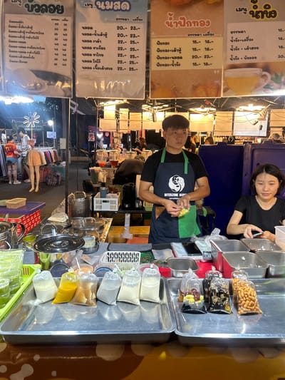 A street food stall operates in Tha Khlong Town Municipality, Thailand, under a string of bright, warm lights at night. A male vendor, wearing a black t-shirt with a green apron, holds a clear plastic bag filled with yellow corn kernels. He stands behind a stainless steel counter laden with various ingredients in clear plastic bags, including white liquids, yellow liquids, dark liquids with toppings, and crunchy snacks. Next to him, a female vendor, wearing a black t-shirt, looks down at her smartphone while preparing items in stainless steel containers. The stall displays three large laminated menus in Thai. The menu on the far left is titled "Bua Loy" (Thai dessert), the middle menu is titled "Nam Misut" (Fresh Milk), and the menu on the right is titled "Nam Fak Thong" (Pumpkin Juice). Behind the vendors, other stalls are visible, with shoppers browsing at one on the left. The overall environment is that of a bustling night market.