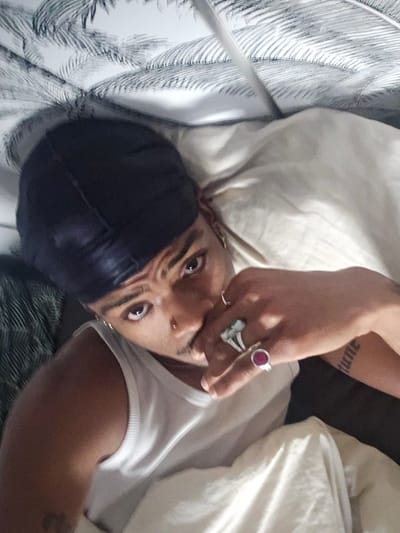 A person is lying down, seemingly on a bed covered with patterned linens. They are wearing a white tank top and a dark-colored durag on their head. The person has multiple piercings including earrings, a nose piercing, and possibly eyebrow piercings. They are wearing rings on their fingers and have a tattoo visible on their arm. The location is unknown and the city of Pantin, France is not discernible from the image.