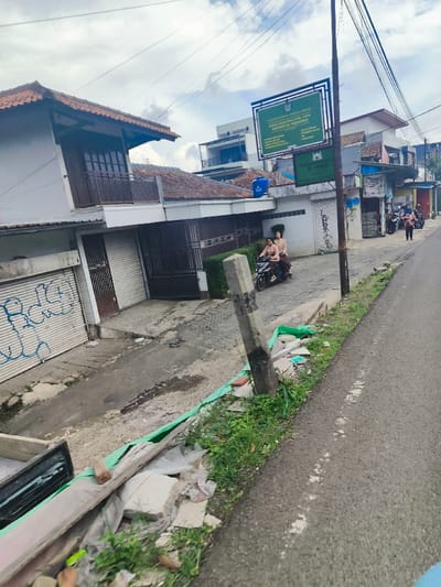 The image shows a street scene in Lembang, Indonesia, at the coordinates -6.83 latitude and 107.607 longitude. The scene features a paved road, a concrete curb, a concrete pole, and a green tarpaulin partially covering a pile of construction debris. The debris includes broken concrete blocks, wood planks, and plastic bags. The buildings on the left side of the road have concrete walls, a red tile roof, and large windows. A blue water tank is visible on the second building. The ground floor of the building on the left is painted white and has a large garage door painted with blue graffiti. The building on the right has a white painted facade and has a small shop on the ground floor with an awning and a large sign hanging above the doorway. On the left side of the image, two women are sitting on a black motorcycle. The motorcycle is parked in the street, and the women are wearing brown dresses and white t-shirts. The background is a cloudy sky. The image was likely captured in a low-traffic residential area.