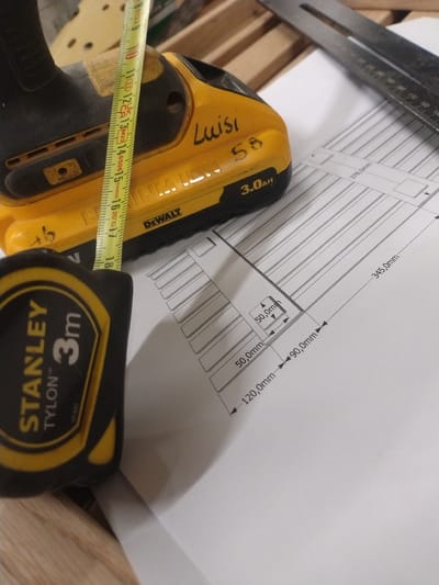 The image depicts a close-up view of a workbench, likely in a workshop or garage, with tools and a technical drawing laid out. A yellow and black DeWalt cordless drill is partially visible in the upper left. A yellow Stanley tape measure, with "3m" clearly marked, is positioned in the lower left corner. A retractable metal measuring tape is laid across the drill, showing metric markings up to 18 cm. In the center, handwritten text "Luisi 58" is visible on the drill. A technical drawing is spread out in the lower right quadrant, featuring dimensions such as "378.0mm", "345.0mm", "50.0mm", "90.0mm", and "120.0mm". A metal L-square ruler is visible in the upper right corner. The background consists of wooden planks. There is no indication of the city of Leudelange, Luxembourg, within the image itself. The scene suggests a DIY or construction project in progress.