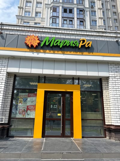 The image shows the exterior of a "Мария-Ра" (Maria-Ra) supermarket in Barnaul, Russia. The building is modern with a light-colored brick facade and large glass windows. Above the entrance, the "Мария-Ра" logo is prominently displayed with a stylized sun and green lettering. Inside the store, visible through the windows, are shelves stocked with merchandise and promotional posters. The entrance features double glass doors framed by a bright yellow structure. On the door, a sign displays the store's hours: 8-22. A person, likely a shopper, is reflected in the glass door. The area in front of the store is paved with dark gray tiles, and a small set of steps leads up to the entrance. The sky above is partly cloudy.