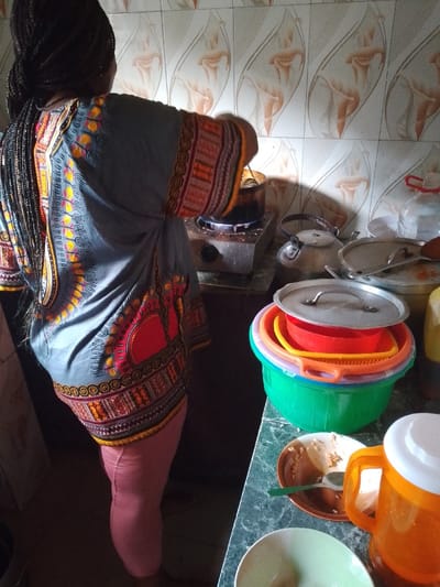 This image captures a domestic kitchen scene in Mgbuoshimini, Nigeria, focusing on a person engaged in cooking. The main subject is a woman, seen from behind, with her back to the camera. She has dark, braided hair pulled back. She is wearing a loose-fitting, short-sleeved top adorned with a vibrant, intricate geometric pattern in shades of grey, red, orange, yellow, and black. Her lower body is clad in pink-colored leggings.

The woman is standing in front of a single-burner gas cooker, which has a pot on it over an active flame. Her right arm is extended towards the pot, suggesting she is stirring or tending to its contents.

To the right of the cooking area, a kitchen counter is visible. It holds a variety of kitchen items, indicating a functional and busy space. These include a stack of nested colorful plastic bowls and colanders (green, red, yellow, pink) with a metal pot lid resting on top. Beside them, a metal kettle and other pots are visible. Closer to the foreground on the counter, there are dirty dishes, including a brown plate with food remnants and a spoon, alongside an orange pitcher with a white lid and a light green bowl. The countertop itself is mottled green and white. The wall behind the cooking area and counter is covered with light-colored tiles featuring a subtle, repeating patterned design. The floor is dark.

The lighting suggests it is daytime, with no strong shadows indicating direct sunlight. No visible text is present in the image. The scene conveys a moment of everyday activity within a home kitchen.