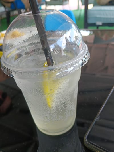 A close-up shot shows a clear plastic cup filled with a fizzy, pale yellow beverage, likely lemonade, with ice and a lemon wedge. A dark straw with a visible portion of a yellow object submerged in the drink. The cup is covered with a clear dome lid, on which water droplets have formed. The cup rests on a reflective dark surface, possibly a table. In the background, out of focus, are hints of outdoor seating, with glimpses of blue and green. The scene is consistent with a beverage being served at an outdoor cafe or restaurant in Harmanli, Bulgaria.