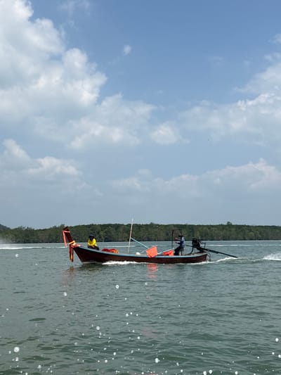 A traditional Thai longtail boat traverses a body of green-blue water near Ko Panyi, Thailand, under a partly cloudy sky. The boat, with a dark brown hull, light blue trim, and red ornamental structures at its bow, is actively moving from left to right, generating a white wake and spray from its stern. A person wearing a yellow shirt and dark head covering is seated in the bow section. Towards the stern, a second individual in a dark shirt and hat stands, operating the boat's longtail engine, which extends into the water. Inside the boat, orange objects, possibly buoys, and red netting are visible. A slender pole rises diagonally from the boat's mid-section. Numerous small, reflective bubbles dot the water surface in the immediate foreground. The background features a distant coastline densely covered with green vegetation, likely mangrove forests, beneath a bright sky with scattered white cumulus clouds.