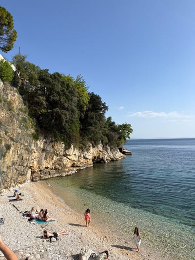 The image captures a vibrant, sun-drenched scene at a pebbly beach in Grad Rijeka, Croatia. The clear blue sky dominates the upper portion, meeting a calm, turquoise sea that stretches towards the horizon on the right. A prominent, rugged cliff face, heavily adorned with lush green trees and vegetation, anchors the left side of the frame, descending to the beach. Evidence of rockfall netting is visible on parts of the cliff, suggesting maintenance for safety.
Several individuals are present on the beach, engaged in leisure activities. A woman in a white dress stands near the water's edge on the right, looking out at the tranquil sea. Closer to the base of the cliff, another woman in a red-patterned dress lies relaxed on the pebbles, appearing to sunbathe, with a green object (possibly a fruit or small item) nearby. In the lower-left section of the beach, a small group of people are gathered, resting on towels and mats, further enjoying the sunny weather. One person lies face down, while others are seen sitting or reclining, with a dark bag visible beside them.
The water is exceptionally clear near the shore, allowing the light-colored pebbles on the seabed to be distinctly visible. A small wooden structure, possibly a pier or platform, extends from the cliff into the sea. The bright sunlight suggests it is mid-day or early afternoon, contributing to the overall atmosphere of relaxation and natural beauty. No visible text is present in the image.