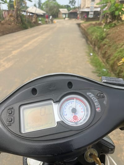 The image depicts a first-person view from a stationary Kymco scooter located in Ikot Ekpene, Nigeria, under an overcast sky. The scooter's dashboard is prominent in the foreground, displaying a digital speedometer reading "0 Km/h," a time of "PM 5:13," and an odometer value of "26384." An analog tachometer indicates 0 RPM, with an illuminated battery warning light. A brass-colored key is inserted into the ignition.

The view extends over an unpaved, light-brown dirt road that recedes into the distance. Both sides of the road are lined with green vegetation, including grass and various trees, some resembling banana plants. On the left, several simple structures with corrugated metal roofs are visible among the trees and exposed reddish-brown soil. Two indistinct human figures are discernible further down the road on the left, one appearing to wear a red shirt. On the right side of the road, a vegetated drainage ditch runs parallel to the path. Beyond the ditch, more foliage and distant buildings are visible, including a multi-story light-colored concrete structure. A faint yellow object, possibly a container, is situated near the road's right edge in the background.