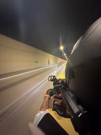 A first-person perspective shot captures a motorcycle traveling through a tunnel at night. The tunnel walls are a light beige color, illuminated by overhead lights that create streaks of light due to motion blur. A white line marks the edge of the road. In the foreground, the rider's gloved hands are visible on the handlebars, with an orange watch on the left wrist. A tattoo is visible on the rider's arm. The motorcycle's windshield and part of the rider's helmet are prominent. A rear-view mirror reflects the tunnel lights. The overall impression is one of speed and movement through an urban or suburban infrastructure. The location is identified as a tunnel in Đurmani, Montenegro.