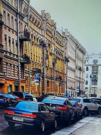 The image depicts a street scene in Naberezhnye Chelny, Russia, characterized by multi-story buildings with ornate architectural details, likely in a historical or classical style. The buildings feature numerous windows, balconies, and decorative elements such as arched openings and cornices. A blue parking sign with a white 'P' is visible on a pole to the right of the street. The street itself is lined with parked cars, suggesting a typical urban environment. Among the vehicles are a dark-colored sedan in the foreground with its taillights illuminated, and other cars parked further down the street, including a white SUV. The sky is overcast, indicating a cloudy day. The overall impression is that of a cityscape with classic architecture and active street life.