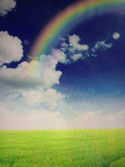 The image depicts a vibrant, digitally rendered scene of a lush green field stretching to the horizon under a bright blue sky. A prominent rainbow arches across the upper portion of the sky, with fluffy white clouds scattered around it. The foreground is dominated by the verdant field, appearing soft and slightly blurred, suggesting a natural landscape. The sky transitions from a deep blue at the top to a lighter hue near the horizon. There are no people or man-made structures visible in the image, suggesting an idealized or naturalistic depiction of the environment. This scene does not depict the city of Qorowul, Uzbekistan, or any recognizable urban or specific geographical location.