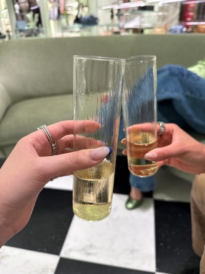 Two individuals are holding sleek, textured champagne flutes filled with a golden-yellow liquid, likely sparkling wine or champagne, in an indoor setting in Zapopan, Mexico. The shot is taken from a slightly overhead perspective, showing only their hands and portions of their bodies, suggesting a celebratory or social moment, possibly a toast.

One hand, positioned higher, features manicured nails with a natural pinkish hue and a silver ring with circular motifs on the middle finger. This person is wearing blue jeans, and their foot in a green shoe is visible on the floor in the background. The other hand, lower in the frame, also has manicured nails, with the index finger adorned with a blue tip. This hand wears two silver rings: one with a braided or twisted design on the ring finger and another, partially visible, with small indentations on the middle finger.

The glasses themselves are clear with a distinct horizontal textured pattern. The background consists of a stylish environment with a prominent black and white checkerboard floor. A soft green or grey upholstered sofa or seating is visible, along with blurry display shelves to the left, which contain various items suggesting a retail or lounge space. No discernible text is visible in the image.