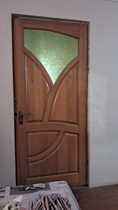 A wooden interior door with a patterned green glass insert at the top stands against a light-colored wall. The door has carved detailing and a metal door handle. A key is visible in a lock mechanism on the left side of the door. In the foreground, a table or surface with a patterned cloth is partially visible, displaying an arrangement of objects that appear to be twigs or sticks. The floor is wooden and appears worn. The scene is indoors, likely in a residential setting in Krynychky, Ukraine.