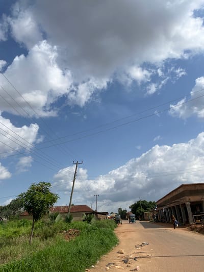 The image captures a street scene in Udu, Nigeria, under a partly cloudy sky. The road is unpaved and appears dusty, with scattered litter along its edges. On the left side of the road, there is overgrown green grass and a small tree. Further back, several buildings with corrugated iron roofs are visible, along with utility poles and electrical wires crisscrossing the sky. On the right side, a building under construction or with an unfinished facade features a row of columns. Several people are present in the scene; one individual is walking on the right side of the road, carrying items, and two individuals are seen near the buildings. A few small vehicles, possibly auto-rickshaws, are present on the road. The overall environment suggests a semi-urban or rural setting within Udu.