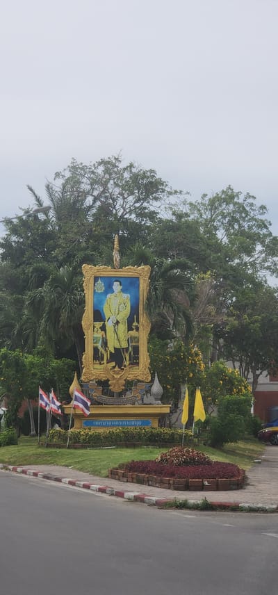 The image depicts a portrait of a man in ceremonial attire, likely a king, displayed in an ornate gold frame. The portrait is positioned outdoors, surrounded by lush green foliage and trees, with a bright blue sky and cloudy haze suggesting daytime. There are yellow flags planted in the ground near the portrait, and three Thai flags are visible in the foreground.
The portrait itself features a man wearing a yellow, intricately patterned outfit and a golden crown. He is seated, with his hands resting on his lap. Behind him, there appear to be other figures, possibly attendants or members of royalty, also in ceremonial dress. The frame is elaborate, with decorative carvings and a gilded finish.
The scene is set in a public area, likely a roadside or park, given the presence of a paved road and a sidewalk with decorative brickwork and plants. The overall atmosphere appears respectful and solemn, fitting for a portrait of a dignitary. The location cues, such as the Thai flags and the script on a blue sign behind the portrait, strongly indicate that this is in Thailand, specifically Nathon. The text on the blue sign is in Thai script and appears to be a place name or official designation. The presence of trees, green grass, and flowering bushes suggests a tropical climate. The lighting implies it is a bright day, possibly overcast due to the haze.