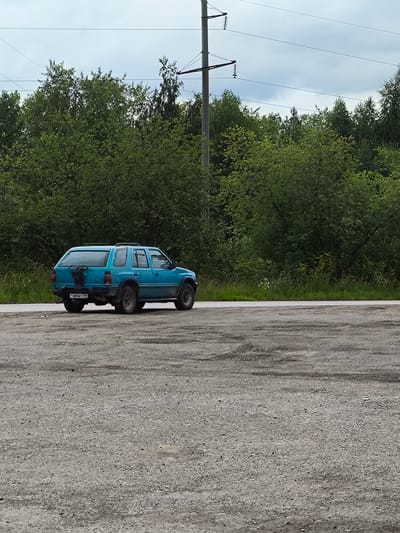 A teal-colored, older model SUV is parked on a gravel shoulder next to a paved road. The vehicle is viewed from the rear passenger side. The SUV has a spare tire mounted on the tailgate. The license plate is visible and reads "T 480 BY 109". The vehicle is surrounded by lush green trees and foliage, suggesting a rural or semi-rural setting. A power line pole with overhead wires runs parallel to the road in the background, with a cloudy sky above. The ground is covered with small stones and dirt, with tire tracks visible in places. The scene appears to be an outdoor location in Solikamsk, Russia.