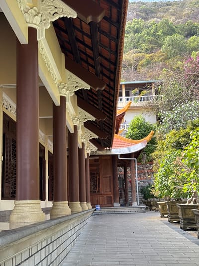 The image displays a long, covered outdoor walkway or veranda, seen from a slightly elevated perspective, looking forward along its length. The photo appears to have been taken in portrait mode and is rotated 90 degrees clockwise.
In the immediate foreground, to the left (which would be the top if rotated correctly), is the underside of a traditional tiled roof or overhang. Dark wooden beams form a grid pattern, supporting reddish-brown roof tiles. Beneath this roof structure, a series of light-colored, decorative corbels or brackets support the eaves. These corbels feature intricate, carved floral or organic motifs in white or very light yellow.
Below these decorative brackets, a series of thick, round pillars run parallel to the view, extending into the distance. These pillars are a dark reddish-brown color, likely wood or painted to resemble wood. They are evenly spaced, creating a rhythmic repetition. The base of each pillar, where it meets the ground level of the walkway, is also light-colored and appears to have some decorative carving.
The walkway or floor itself, running alongside the pillars, is made of light grey square tiles, laid in a grid pattern. To the right of the pillars, further into the scene, a slightly darker, checkered patterned surface is visible, possibly another type of paving or a drain.
In the mid-ground and background, behind the pillars and walkway, a lush natural landscape with dense green trees and foliage is visible. Some parts of the trees show lighter green leaves, suggesting new growth, and a few hints of purple or pink flowers can be seen among the greenery. Beyond the trees, what appears to be part of another building or structure with a lighter-colored wall is visible.
Towards the right side of the image, closer to the background, there's another section of a building with a curved, upturned roof typical of traditional Asian architecture. This section also features dark wooden elements and intricate carvings on what might be a doorway or window frame. A system of pipes, possibly for drainage or utilities, runs along the wall of this building. There appears to be a small blue sign with white text on it near the pipes, but the text is not legible.
The lighting suggests it is daytime, with bright sunlight creating some highlights and shadows, particularly on the roof tiles and the foliage. The overall impression is one of a serene, possibly temple or traditional garden setting, characterized by detailed craftsmanship and natural beauty.