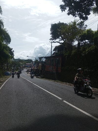 The image depicts a daytime scene on an asphalt road in Sandamal Eliya, Sri Lanka, under an overcast sky with white and grey clouds. The road, featuring a solid white centerline, extends from the foreground into the midground, curving slightly to the left.
In the immediate foreground on the right, a white and red scooter is traveling leftward, carrying two occupants. The driver is wearing a light brown jacket and a dark helmet, while the passenger behind them is partially obscured.
Along the right side of the road, a two-story building is visible. Its lower level is reddish-brown with rectangular windows, and an external dark metal staircase ascends to the upper level, which features a white facade and a dark roof. Utility poles with multiple overhead power lines are positioned near the building. Directly behind and above this structure, a steep embankment is covered in dense green vegetation and large trees.
Further down the road in the midground, multiple motorcycles and scooters are visible, traveling away from the viewer. One distinct light-colored scooter with a sidecar attached is present among them on the right side of the road. In the distance, other vehicles and buildings are discernible, indicating a populated area.
On the left side of the road, a thick wall of dense green foliage, consistent with bamboo or similar tall plants, borders the roadway. The ground immediately adjacent to the road's edge on the right is unpaved and rocky.