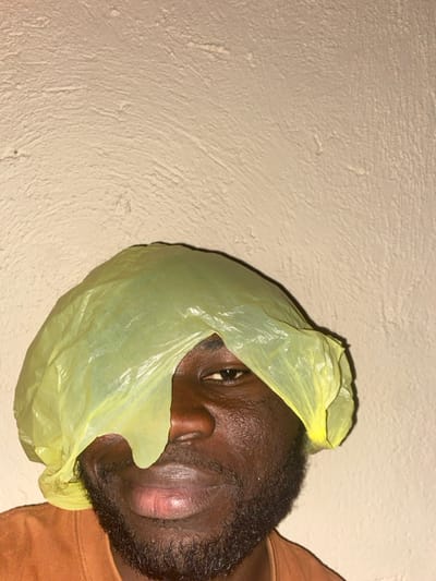 An adult male with dark skin and a beard is depicted in a medium close-up, centered against a light beige, textured wall in Udu, Nigeria. He is wearing a translucent, crinkled yellow plastic bag on his head, which drapes over his forehead and the left side of his face, partially obscuring his left eye and the bridge of his nose. His right eye is visible and directed towards the camera. His mouth is closed. He is wearing a brown or terracotta-colored garment, visible at the bottom of the frame. The lighting source is not directly visible, but illuminates the subject and wall, revealing surface textures.