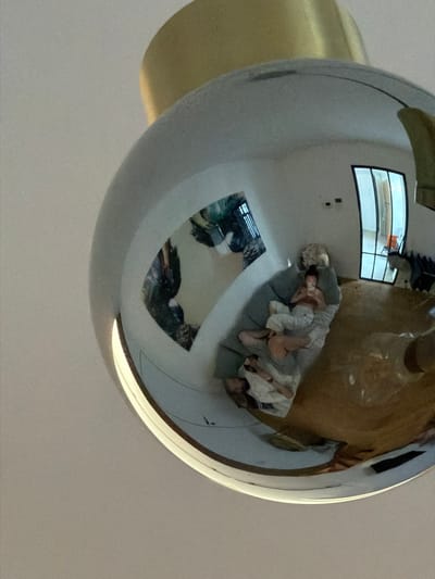 This fisheye reflection captures an interior scene, likely a living space. At the center of the reflection, two people are visible, nestled together on a sofa. One person is lying on their side, facing away from the viewer, while the other is partially curled up against them. They appear to be in pajamas or comfortable loungewear. The reflection also shows a window with multiple panes in the background, suggesting a modern interior. The overall mood is intimate and relaxed, with the soft lighting indicating it could be daytime or evening. The reflection on the spherical object, likely a decorative orb, distorts the scene, creating a wide-angle perspective. The metallic surface of the orb also reflects a golden-toned object, possibly a lamp or part of the room's decor, and a portion of the ceiling. The context of Bruges, Belgium, does not provide specific visual cues within this reflection.