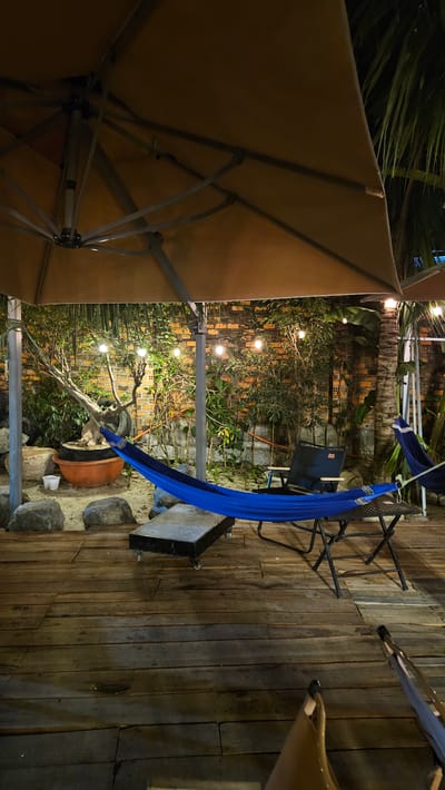 This media file captures an outdoor patio or garden area at night, likely in Cam Đức, Vietnam. The scene features a cozy and relaxed atmosphere, illuminated by warm string lights.
In the foreground on the left, a large, beige patio umbrella extends, with its fabric and support structure visible from below. To the right of the umbrella, a bright blue fabric hammock is suspended, appearing ready for use. Adjacent to the hammock is a dark, foldable outdoor chair.
The background reveals a rustic brick wall partially covered by lush green foliage, including various plants and what appears to be palm fronds. The string lights are integrated into the greenery, casting a soft glow. On the ground, there is a wooden deck floor, and below the hammock, a large terracotta pot holds a sculptural plant or bonsai-like tree, surrounded by several rocks.
The image conveys a tranquil evening setting, perfect for relaxation. No people are visible, and there is no discernible text in the frame.