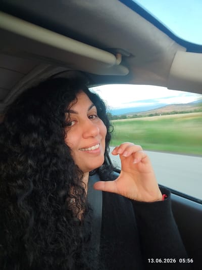 A woman with long, dark, curly hair is captured in a selfie from inside a vehicle. She is smiling at the camera and making a heart shape with her fingers. The background shows a blurred landscape of green fields and hills under a bright sky. The timestamp "13.06.2026 05:56" is visible in the lower right corner of the image. The scene is likely a road trip given the motion blur of the landscape.