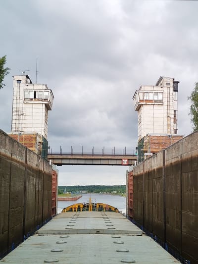 The image shows a view from the deck of a cargo ship, looking forward through a lock chamber towards the outside. Two identical, weathered, white concrete control towers stand on either side of the lock. These towers have multiple windows and access ladders. A metal bridge connects the tops of the towers, with a red sign displaying "15.9" visible on its underside.
The lock walls are tall and made of large, dark concrete sections, showing signs of age and water staining. The ship's deck, made of grey, flat metal panels with numerous circular hatches, occupies the foreground and extends into the lock. The ship itself is a cargo vessel, with a yellow superstructure and cabin visible further forward.
Beyond the lock, the water of a river or canal is visible, with a distant tree-lined shore and some buildings on the horizon. A large industrial crane can be seen in the distance on the right. The sky is overcast with heavy grey clouds. The location is identified as the Vytegra Lock on the White Sea-Baltic Canal in Vytegra, Russia. No people are visible in the image.