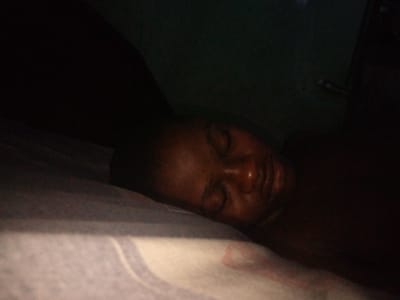 A person's face is partially visible in the foreground, resting on a pillow. The lighting is dim, suggesting an indoor setting, possibly a bedroom. The background is dark and indistinct. The image is composed from a close-up perspective, focusing on the individual's features. The scene appears to be in Shangisha, Nigeria.