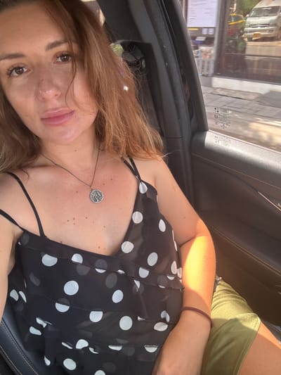 A woman with auburn hair and light skin is pictured from the chest up, sitting in the driver's seat of a car. She is wearing a black and white polka dot top and a silver pendant necklace. The interior of the car is visible, with the door frame on the right and the window on the left. Through the window, a street scene in Baan Bang Rak, Thailand, is visible, with a white van and a yellow vehicle, possibly a tuk-tuk, driving by. Sunlight streams into the car, casting shadows on the woman's face and arm.