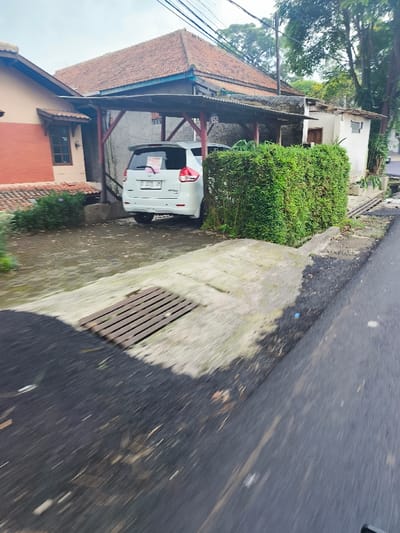 The image shows a white car parked under a carport in front of a residential building in Lembang, Indonesia, located at coordinates -6.822 latitude and 107.61 longitude. The car is parked on a concrete driveway, and there is a dense, green hedge separating the driveway from a paved road. There is a drain cover on the driveway in front of the hedge. The road is paved and asphalt, and the edges of the road are covered with leaves and debris. The residential building appears to be a two-story structure with a red tile roof and a wooden carport. There is a small garden area in front of the building with some greenery. The scene is taken from a perspective of a vehicle driving on the paved road, providing a wide shot of the car and the surroundings.