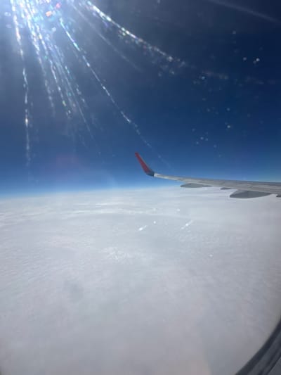 The image shows an aerial view from an airplane window, looking out at the Earth's curvature and the sky. The perspective is rotated, with the airplane's wing prominently featured on the right side of the frame. The wing is a light gray color with visible struts and the tip is painted red. Below the wing, a vast expanse of white clouds blankets the Earth's surface, creating a textured, undulating pattern. To the left of the clouds, the sky transitions into a deep, vibrant blue, indicating a high altitude.

Scattered across the blue sky are numerous bright, bokeh-like orbs of light, as well as streaks of light that appear to be caused by reflections or light refracting through the airplane window. These light effects are particularly concentrated in the lower left portion of the image, suggesting the sun is in that direction and its rays are interacting with the window's surface. There are no discernible people or specific objects other than the airplane wing.

The scene suggests an ongoing flight, likely during daytime, given the bright sunlight and clear blue sky. The thick cloud cover below implies a substantial altitude, obscuring any ground-level details or specific location cues, though the provided context suggests Zhukatau, Russia. The overall impression is one of vastness, altitude, and the visual phenomena associated with air travel.