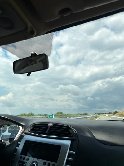 The image is a first-person view from inside a car driving on a highway, likely in or near Aleksandrovo, Bulgaria, under a cloudy sky. The dashboard of the car is visible in the foreground, with the steering wheel on the left and a section of the car's console with a radio. A rearview mirror is hanging from the windshield. Outside the car, the highway stretches ahead, with a speed limit sign indicating "52" visible on the right. The surrounding landscape appears rural, with fields and trees lining the road. A few other cars are present on the highway in the distance. The overall scene depicts a journey on a highway on a day with mixed cloud cover.