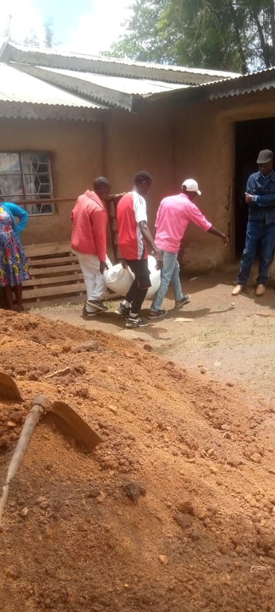 The image shows four men carrying a large white sack in front of a building with a corrugated metal roof, located in Burundu, Kenya, at coordinates 0.46 latitude and 34.776 longitude. The men are wearing casual clothing. One man is wearing a red shirt, another a pink shirt, another a white shirt, and the last man is wearing a red jacket. There is a woman standing in the background, behind the men, wearing a floral dress. The ground is covered in loose, brown dirt and there is a shovel lying on the ground in the bottom left corner of the image. There is a wooden pallet to the left of the men. The scene appears to be taking place in the yard of a residential property. The building is constructed with concrete and has a dirt floor. The scene is bright and the weather is clear.