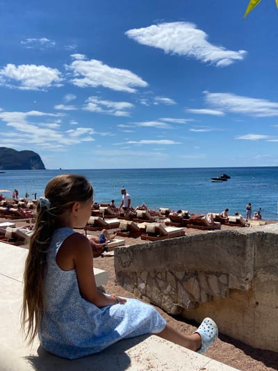 In Petrovac centar, Montenegro, a young girl with long, brown hair tied back with a blue scrunchie, wearing a blue floral dress and a blue striped croc shoe, sits facing away from the camera. She is positioned on a concrete ledge overlooking a beach scene. The beach is lined with rows of beige lounge chairs with umbrellas. The blue ocean stretches out to the horizon, where a large, dark green hill or mountain is visible on the left. Several people are scattered across the beach, some lounging on chairs, others in the water, and one man in a white shirt is walking. Two small boats are present in the water. The sky above is a vibrant blue, dotted with white, fluffy clouds. A rough, stone wall is in the foreground, partially obscuring the view of the beach.