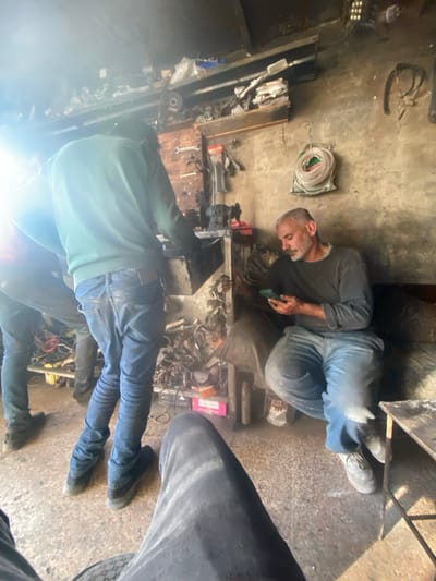 This image displays an interior scene within a workshop or mechanical repair space in Ma'raba, Syria. Four individuals are present, three of whom are clearly visible. In the immediate foreground, the legs of an individual wearing dark trousers are centrally positioned. To the left, an adult male wearing a light-colored long-sleeved sweater and blue jeans stands with his back mostly to the viewer, bent over a dark workbench. A second male, partially visible to the left of the first, also stands near the workbench, wearing dark trousers. On the right, a third adult male with short grey hair and a beard, dressed in a dark grey long-sleeved shirt and blue jeans, is seated on a low, dark-colored bench-like structure. He holds a smartphone in both hands and is looking down at its screen.

The environment features light-colored, possibly concrete walls. Elevated shelves above the workbench are cluttered with various tools, metal components, plastic bags, and other miscellaneous items. A coiled white cable or hose hangs on the right wall. The workbench area contains tools and what appears to be small machinery. The floor is a rough, light-colored concrete surface, with some debris and objects scattered. Bright sunlight enters from an unseen source in the upper left, causing significant lens flare across the upper-left quadrant of the image.