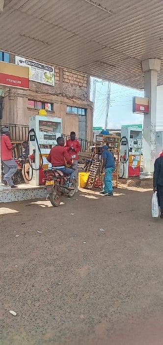 The image captures a bustling scene at a fuel station in Kipkarren, Kenya, during what appears to be a bright, sunny daytime. The overall setting is somewhat informal, with unpaved ground and basic structures.

Several individuals are present, engaged in various activities related to fueling. In the mid-ground, a man dressed in blue work attire and a cap stands near a set of fuel pumps, seemingly attending to a transaction or filling containers, with a large yellow container visible beside him. Closer to the foreground, two men, both wearing red tops, are seated on a motorized tricycle or cart. One appears to be operating the vehicle, while the other is a passenger. The vehicle is positioned near another fuel pump, suggesting they are waiting to be served or have just been fueled. Another person in a red top and grey pants is partially visible, kneeling or bending near a different pump in the foreground. In the upper right background, a person in dark clothing carrying a white bag is also visible, perhaps walking by or waiting.

The fuel station features multiple white and green fuel pumps. Above the pumps, there is a corrugated metal canopy providing shade. Behind the pumps, a building with exposed brickwork and windows is visible. The ground is a rough, dusty, unpaved surface.

Visible text includes "PREMIUM" on a red and gold sign, and partially legible text on another sign reading "ONE MOTOTRCYCLE" and "CHEMOTIONC'S CHEAP." The scene depicts typical daily life and commerce at a local fueling point in the region.