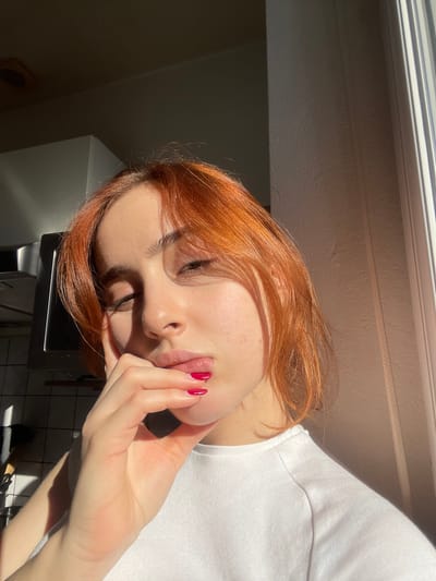 The image depicts a young adult female situated indoors in Reims, France. She possesses short, reddish-orange hair and fair skin. Her left hand, with fingers curled and nails painted bright red, rests against her chin and mouth. She is wearing a plain white t-shirt. Her eyes are partially closed, appearing to squint. Strong, direct sunlight enters from a visible window on the right side of the frame, illuminating the subject's face and creating harsh shadows. The background on the left reveals elements consistent with a domestic kitchen, including a dark metallic range hood and a light-colored tiled backsplash, largely obscured by shadow.