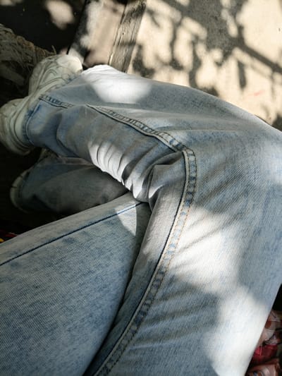 The image shows a close-up view of a person wearing light blue, acid-washed denim jeans and white sneakers. The person is seated outdoors, with sunlight casting shadows on their legs and the ground. The ground appears to be made of dirt or gravel, with some wooden planks visible. The scene is likely a public space within the city of Osh, Kyrgyzstan, possibly a park, street, or courtyard, given the combination of natural elements and man-made structures. There are no other people or significant environmental features visible.