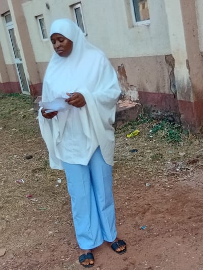 A woman wearing a white head covering and light blue pants stands outside a building. She holds a piece of paper in her hands. The ground is dirt and grass. She wears dark sandals. The building is light-colored with windows. The scene appears to be outdoors during daylight.