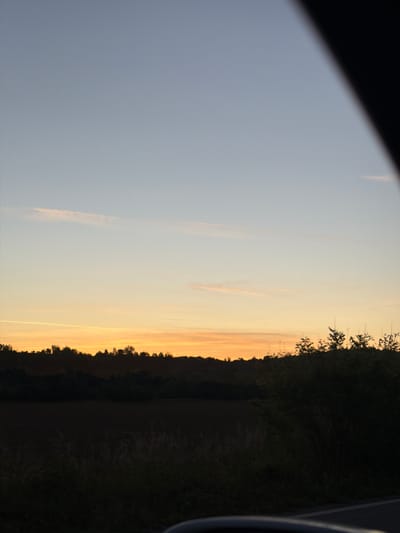 The image depicts a sunset viewed from a vehicle, likely a car. The location is near Novi Chiflik, Bulgaria, evidenced by the landscape which consists of a tree line along the edge of a field. The sky is a gradient of blue and orange as the sun sets. Part of the vehicle frame is visible in the image.