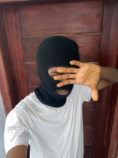 The image displays a single individual in Joma, Ghana, positioned against a background of reddish-brown wooden paneling. The individual is wearing a black knitted balaclava that covers the entire head and face, with openings for the eyes and mouth. A plain white, short-sleeved t-shirt is visible. The person's right hand is raised, palm outwards, with fingers slightly spread, partially obscuring the eye openings of the balaclava. The visible skin on the hand and through the balaclava openings is dark-toned. The wooden background shows visible grain and appears to be composed of vertical planks with horizontal joints.