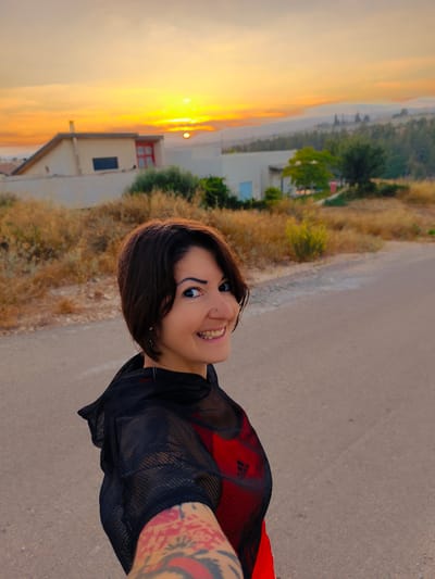 A woman with short dark hair and a nose ring smiles at the camera, her face partially visible. She is wearing a black mesh top over a red athletic shirt, and her left arm, adorned with tattoos, is extended towards the viewer. The woman stands on an asphalt road with dry vegetation on the left and a few buildings in the background. The scene is illuminated by the warm glow of a sunset over a hilly landscape, suggesting a rural or semi-rural setting within the Lachish Regional Council, Israel. The sky is a vibrant mix of orange and yellow hues, with the sun visible near the horizon.