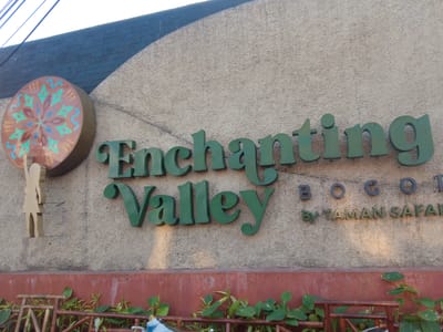 A textured, light-colored wall serves as the backdrop for a sign that reads "Enchanting Valley" in large, dark green serif font. To the left of the text, a circular emblem with a floral design in shades of teal, brown, and yellow is mounted on the wall. Below the emblem, a cutout of a stylized human figure in a light brown color stands. To the right of "Enchanting Valley," the word "BOGOR" is displayed in black block letters, followed by "BY TAMAN SAFARI" in smaller green serif font. The wall is topped by a dark gray brick structure and a hint of the sky. The bottom of the image shows a reddish-brown border with green plants growing in front of it, along with a wooden fence-like structure. The scene appears to be an entrance to a park or attraction.