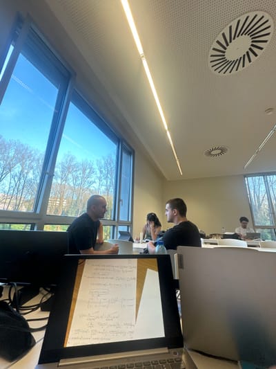 The image depicts an indoor study room or library within the city of Guimarães, Portugal, characterized by natural light from large windows and artificial overhead illumination. The scene is observed from behind an open silver laptop displaying handwritten mathematical equations on its screen, suggesting the viewpoint of someone studying.
In the mid-ground, at a white table positioned against the windows, two adult males are engaged in conversation. The male on the left, with a bald head or very short hair and wearing a black t-shirt, faces right and speaks, holding a white cup. Opposite him, a male with short dark hair, also in a black t-shirt, listens attentively; a blue plastic water bottle rests on the table in front of him. Behind them, further into the room, a female with dark hair is partially visible, focused on a tablet or notebook on her table. In the background, another male with dark, curly hair and glasses is seated at a table, working on a laptop.
The room features a light-colored, perforated acoustic ceiling with multiple linear LED strip lights and circular air diffusers. The large windows reveal a clear blue sky with a faint airplane contrail and numerous leafless deciduous trees outside, indicative of a late winter or early spring season.