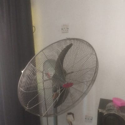 The image depicts an indoor scene in Abakaliki, Nigeria, featuring a large, industrial-style standing fan as the central subject. The fan has a metallic, circular cage guard and dark-colored blades, with a visible red adjustment knob or cap on its central housing. It is angled slightly upwards and towards the left, positioned on a light-colored, possibly white or off-white, textured wall.
On the wall, two electrical outlets are visible: one higher up, appearing to be a two-pin type, and another lower down, which is a three-pin Type G outlet with a white adapter or charger plugged into it. To the left of the fan, a dark grey or black curtain hangs vertically. On the lower right, a dark, indistinct object, possibly a piece of furniture or an electronic device, is partially visible with a pink item resting on its top surface. No people are present, and no specific actions or interactions are depicted within the frame. The environment appears to be a typical room or indoor space.
