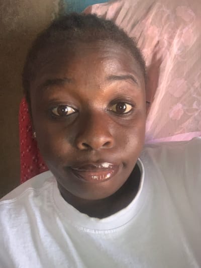 A close-up, high-angle selfie of a young Black woman, likely in her late teens or early twenties, is captured in a room. She is wearing a plain white t-shirt. Her hair is dark and short, pulled back from her face. Her skin is dark brown, and she has light brown eyes. Her lips are glossy and appear to be wearing lip gloss or balm, and she has a slight smile. Behind her, the background is blurred, but a pink, floral-patterned fabric, possibly a sheet or pillowcase, is visible on the right side. On the left, a portion of a red, patterned fabric is also visible. The lighting is soft and appears to be natural light coming from the front. The image is likely taken indoors, possibly in a bedroom or living area, as suggested by the soft furnishings. There is no clear indication of the exact location within Takau, Nigeria, beyond it being an indoor residential setting.