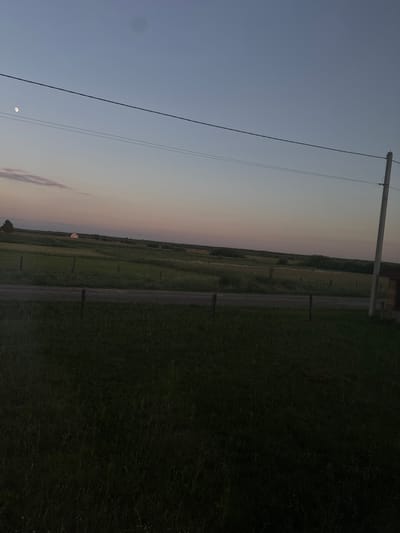 This is a rotated photograph of a rural landscape in Chemerne, Ukraine, likely taken at dusk. The sky transitions from a pale blue at the top to soft hues of pink and orange near the horizon, indicating sunset or sunrise. A utility pole with wires runs diagonally across the image from the upper left to the middle right. In the distance, across a grassy field and a dirt path, a small, light-colored building with a dark roof is visible. Several fence posts dot the landscape. The overall mood is serene and tranquil. The moon is visible in the sky in the lower left quadrant.