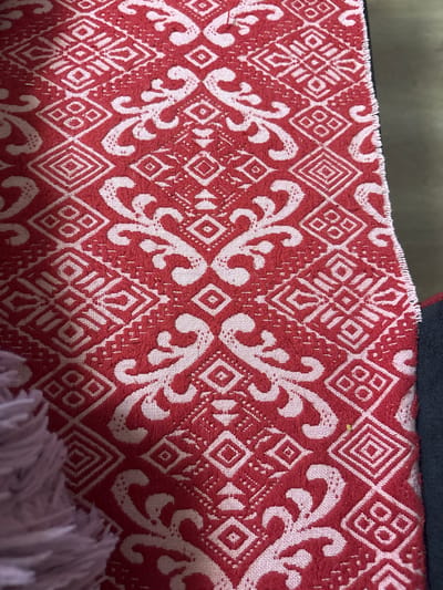 A close-up, top-down view depicts a red and off-white patterned textile, located in the city of Kuklen, Bulgaria. The fabric features an intricate, repeating design composed of prominent off-white stylized floral or scroll motifs interspersed with geometric patterns against a textured red background. The geometric elements include outlined diamonds, some containing concentric squares or lines, as well as distinct square clusters each containing four smaller squares. The red portions of the fabric exhibit a slightly raised, woven or embroidered texture, while the off-white areas appear comparatively flatter. The right edge of the textile displays a tightly woven, fringed, and unfinished seam, indicating a cut edge. A dark, solid-colored material is partially visible adjacent to this edge on the right, and a blurred, light purple/pink fluffy textile is partially visible along the left edge of the frame.