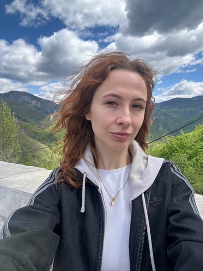 A selfie of a fair-skinned woman with shoulder-length red-brown curly hair. She is wearing a white hoodie under a black Adidas jacket, and a gold necklace. Behind her is a mountainous landscape with trees and vegetation. The sky is partly cloudy with patches of blue. She is in Đurđevića Tara, Montenegro. A gray concrete structure is in the lower right foreground.