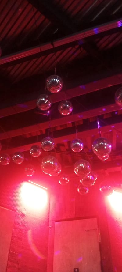 The image shows a dimly lit interior scene, likely a club or event space, with multiple disco balls hanging from the ceiling. The scene is bathed in vibrant red and purple lighting, creating a festive and energetic atmosphere. The disco balls, of various sizes, are reflective and catch the light, scattering it around the room. The ceiling appears to be made of corrugated metal or similar material, with exposed beams. In the lower part of the image, bright lights illuminate a wall or backdrop, possibly a stage or dance floor. The overall impression is one of a lively party or celebration. There is no visible text in the image. The time of day and weather are not discernible, but the setting suggests an indoor event. The location context provided is Tolyatti, Russia.