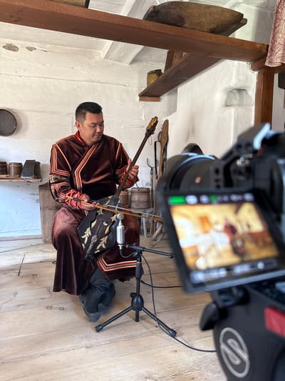In the city of Shushenskoye, Russia, a man is seated indoors, playing a traditional stringed instrument. He is wearing a rich, brown velvet robe with ornate gold and red trim. The instrument, held across his lap, is dark with gold embellishments and has a bow. A microphone is positioned in front of the instrument. The setting appears to be a historical exhibit or museum, with rough white-plastered walls and wooden beams supporting the ceiling. Various rustic artifacts, including wooden barrels and containers, are visible on shelves and on the floor. In the foreground, a camera on a tripod is pointed at the man, with its screen showing a blurred image of the scene. The floor is made of light-colored wooden planks.
