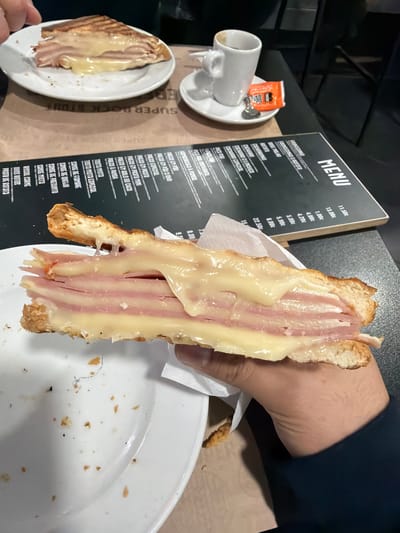 The image captures an indoor dining scene at a commercial establishment in Gondomar, Portugal. In the immediate foreground, a light-skinned hand, partially covered by a dark blue long-sleeved garment, holds a large, open-faced toasted sandwich. The sandwich reveals multiple thick layers of pink processed meat, likely ham, interspersed with substantial amounts of melted yellow cheese that drips from the edges of the golden-brown, crusty bread. A portion of white paper wrapping is visible around the base of the sandwich.
To the left, a white ceramic plate is partially visible, containing various dark and light crumbs, suggesting recent consumption. On the dark, textured tabletop, a brown paper placemat or covering features inverted text, including "SUPER BOCK STOUT." Above this, a black menu board with white text displays a list of items and prices in Portuguese, such as "MENU," "PREGO," "TOSTA MISTA," and "OVOS ROTOS."
Further back on the left, a second white ceramic plate holds a triangular section of a similar toasted sandwich, also covered with melted cheese. To the right, a small white espresso cup sits on a white saucer, accompanied by a silver spoon and an orange "DELTA CAFES" sugar packet. The background is dark and indistinct, with suggestions of other furniture or dark surfaces within the establishment.